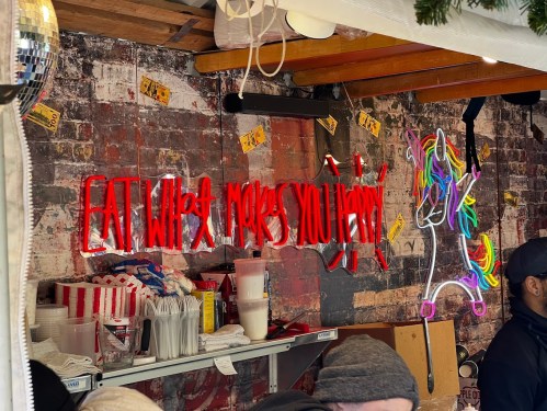 A colorful neon sign reads "EAT WHAT MAKES YOU HAPPY" against a brick wall, with a unicorn illustration nearby.