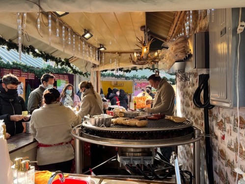 A festive market stall with people in masks, serving food and drinks, decorated with lights and holiday decor.