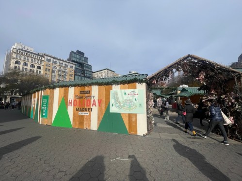 A bustling holiday market with wooden stalls and festive decorations in an urban square, surrounded by people.