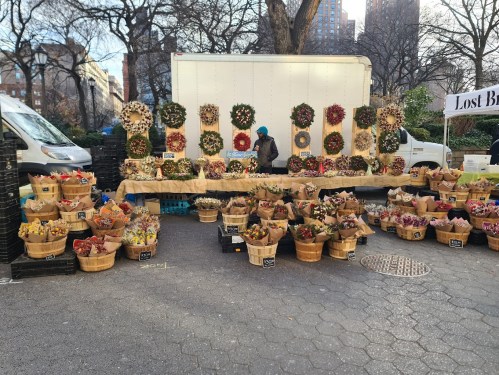 A market stall filled with colorful flower arrangements and wreaths, with a vendor working in a park setting.