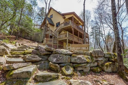 A multi-level house nestled among trees, featuring a wraparound deck and stone steps leading up to it.