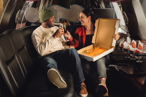 A couple enjoys pizza and wine in a stylish limousine, smiling and sharing a cozy moment together.