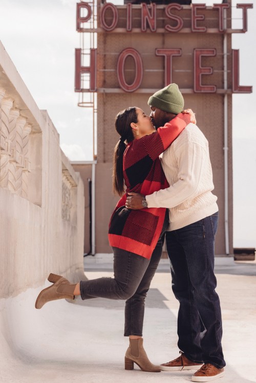 A couple shares a kiss on a rooftop, with the Poinsett Hotel sign in the background. They are dressed warmly and smiling.