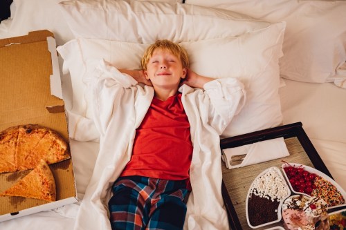 A happy boy in a robe relaxes on a bed, surrounded by pizza and a dessert platter.