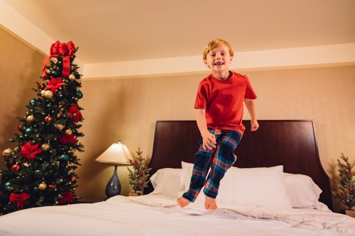A joyful boy in pajamas jumps on a bed, with a decorated Christmas tree in the background.