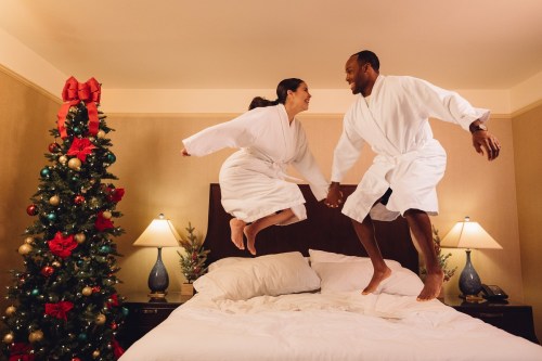 A couple in white robes joyfully jumps on a bed, with a decorated Christmas tree in the background.