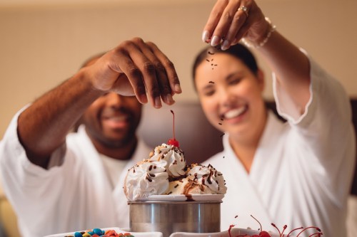 A couple in bathrobes joyfully decorating ice cream sundaes with cherries and sprinkles.