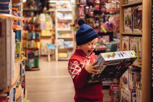 A young boy in a red sweater and blue hat excitedly holds a toy box in a colorful store aisle filled with toys.