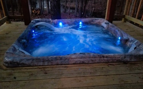 A hot tub with blue lights, surrounded by wooden decking and trees in a serene outdoor setting.