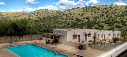 A serene pool area surrounded by mountains and greenery, with a few buildings nearby under a blue sky with clouds.