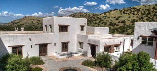 A southwestern-style adobe house surrounded by greenery and hills under a blue sky with fluffy clouds.