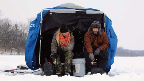 Two people ice fishing in a tent on a snowy lake, bundled up in winter clothing, focused on their fishing lines.