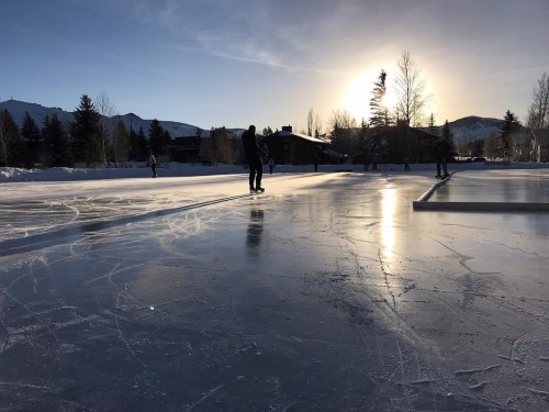 A serene outdoor ice skating rink at sunset, with skaters gliding on the smooth surface and mountains in the background.