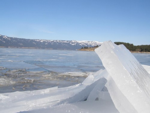 A frozen lake with large ice formations and snow-capped mountains in the background under a clear blue sky.