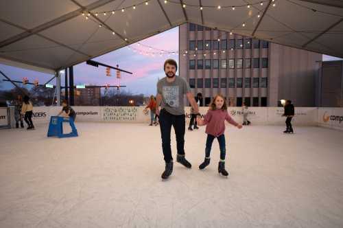 A man and a girl skate together on an ice rink under a tent, with city buildings and lights in the background.