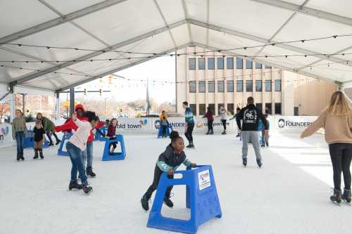 A group of children and adults ice skating indoors, using skating aids, under a large tent.