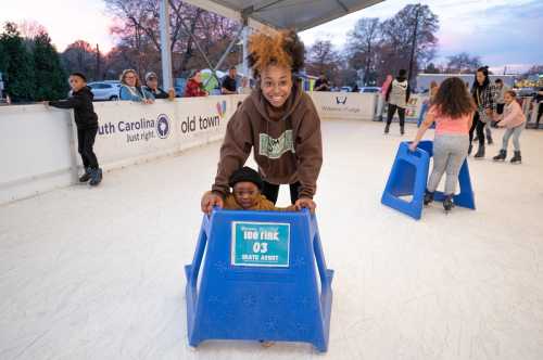 A girl smiles while helping a young child on an ice rink, surrounded by other skaters and a festive atmosphere.