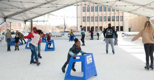 Children and adults ice skating indoors, using blue skating aids, under a large tent with festive decorations.