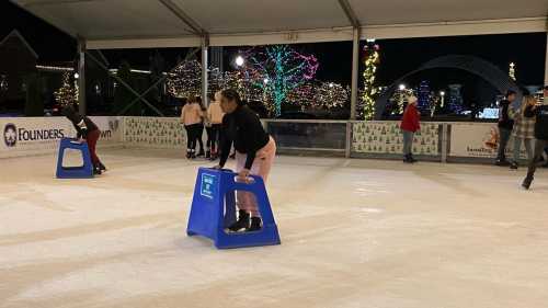 A group of people ice skating at night, using blue skating aids on a rink decorated with colorful lights.