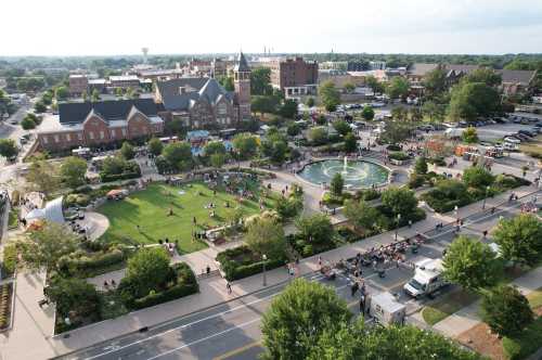 Aerial view of a bustling park with a fountain, people enjoying the outdoors, and nearby buildings in a vibrant town.