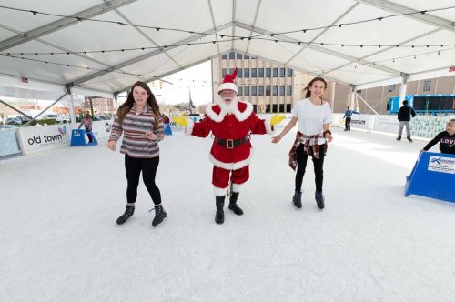 Two girls ice skating with Santa Claus in a festive rink under a tent.
