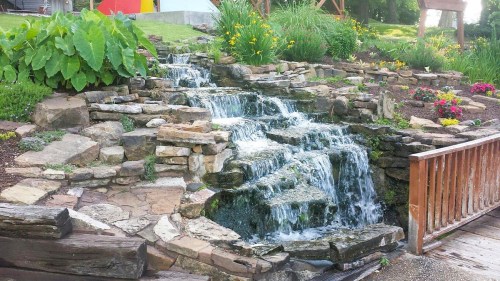 A serene waterfall cascades over rocky steps, surrounded by lush greenery and colorful flowers.