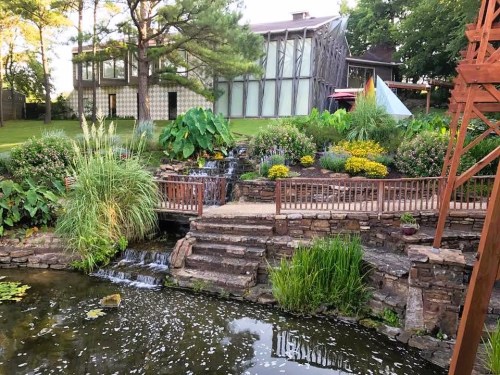 A serene garden with a pond, waterfall, and lush greenery, featuring colorful flowers and a modern building in the background.