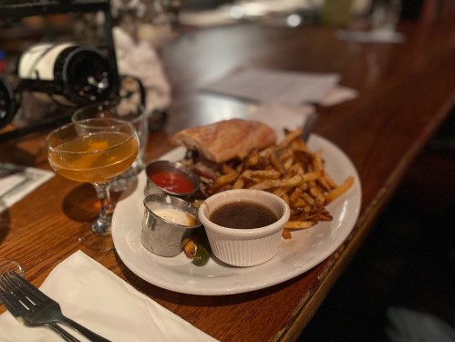 A plate of fries, a sandwich, dipping sauces, and two cocktails on a wooden table in a restaurant setting.