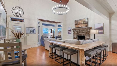 Cozy living room with a stone fireplace, large windows, and a wooden dining table with bar stools. Warm, inviting decor.