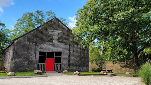 A rustic wooden building with a bright red door, surrounded by trees and a gravel path.