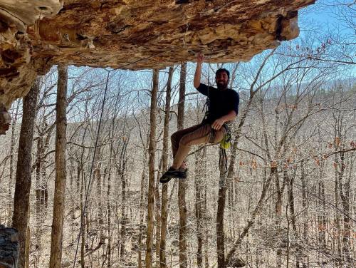 A climber hangs from a rock overhang in a forest, smiling while holding onto a rope. Trees are bare in the background.