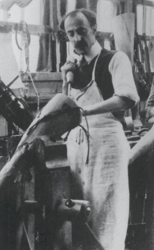 A man in an apron works on a shoe in a workshop, surrounded by tools and materials.