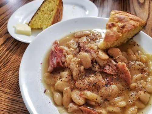 A bowl of white beans with ham, seasoned, served with cornbread and a buttered slice on a plate.