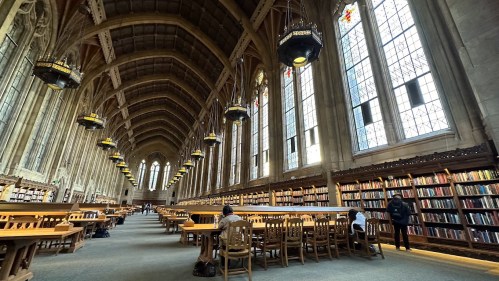 A spacious library with tall windows, wooden tables, and bookshelves, featuring a high ceiling and warm lighting.