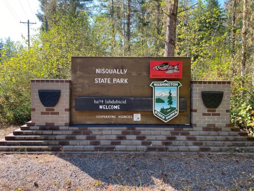 Sign for Nisqually State Park, featuring welcome message and park logo, surrounded by trees and greenery.