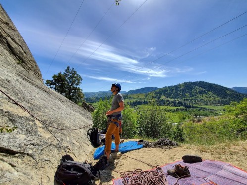 A climber stands near a rock face, surrounded by lush green hills and a clear blue sky, preparing for a climb.