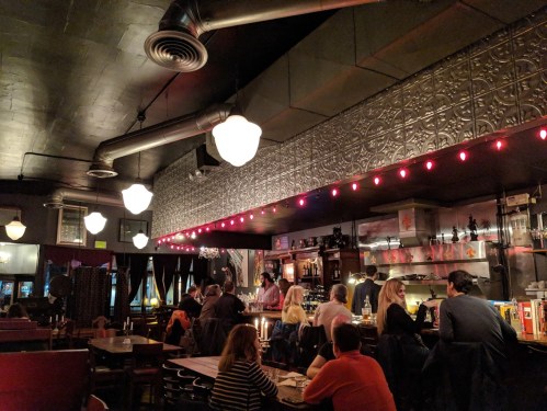 A cozy bar interior with warm lighting, patrons seated at tables, and a bartender serving drinks.