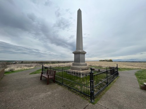 A tall stone obelisk surrounded by a black fence, with a bench nearby, set against a cloudy sky.