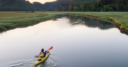 A person paddles a yellow kayak on a calm river surrounded by lush green hills at sunset.