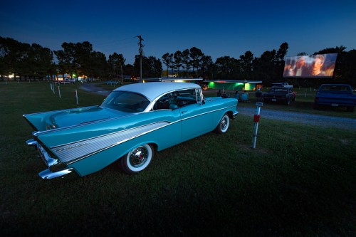 A vintage turquoise car parked at a drive-in movie theater, with a film screen illuminated in the background at dusk.