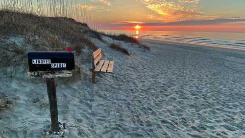 A beach scene at sunset featuring a mailbox labeled "Kindred Spirit" and a wooden bench on the sandy shore.