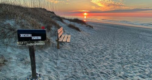 A beach scene at sunset featuring a mailbox labeled "Kindred Spirit" and a wooden bench on the sandy shore.