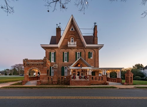A large, historic brick house with green shutters and a decorative porch, set against a twilight sky.