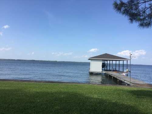 A calm lake scene with a wooden dock and a small white boathouse under a clear blue sky. Green grass in the foreground.