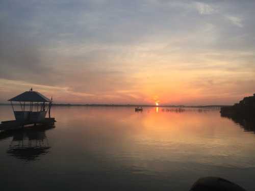 A serene sunset over calm waters, with a small gazebo and silhouettes of distant boats on the horizon.