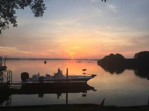 A serene sunset over a calm lake, with a boat docked at the shore and silhouettes of trees in the background.