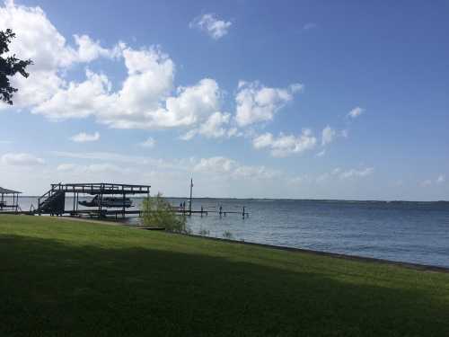 A serene lakeside view with a wooden dock, boats, and fluffy clouds in a blue sky, framed by green grass.