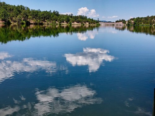 A serene lake reflecting blue skies and fluffy clouds, surrounded by lush green trees and distant hills.