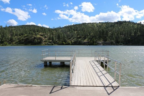A wooden dock extends into a calm lake, surrounded by green hills and a blue sky with fluffy clouds.