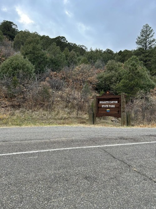 Sign for Sugarite Canyon State Park beside a road, with a backdrop of trees and a cloudy sky.